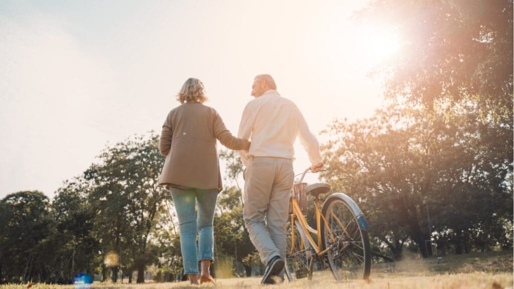 An older couple walking through a park as one pushes a bike